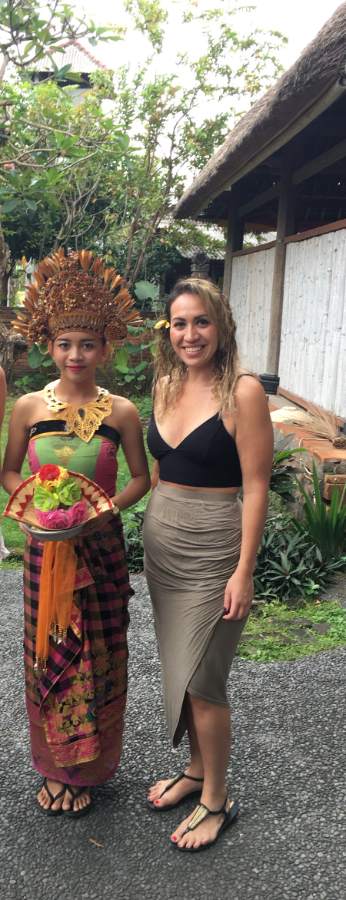 Kiri Kenny smiling in a tropical garden, a flower tucked in her hair, standing beside a Balinese dancer